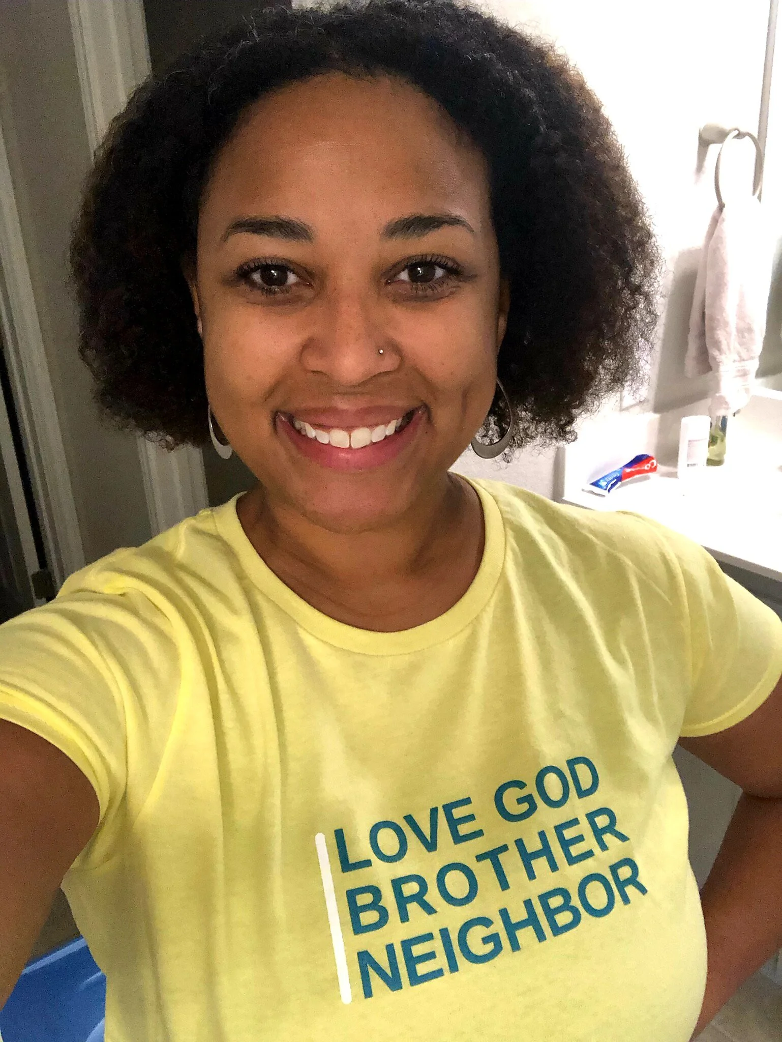 Woman wearing a yellow t-shirt with the text 'LOVE GOD BROTHER NEIGHBOR', smiling in a bathroom.
