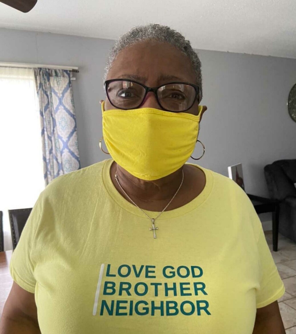Person wearing yellow mask and shirt with text 'LOVE GOD BROTHER NEIGHBOR', standing indoors.