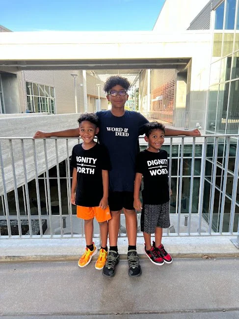 Three kids standing on a walkway, smiling, with two wearing "DIGNITY WORTH" shirts, and one wearing "WORD DEED" shirt, against an urban building backdrop.