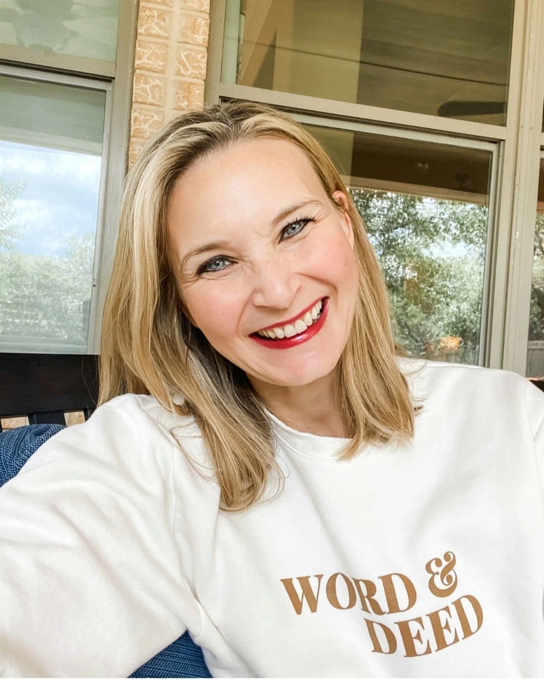 Smiling person wearing a white sweatshirt with the text 'WORD & DEED', sitting outside near large windows.