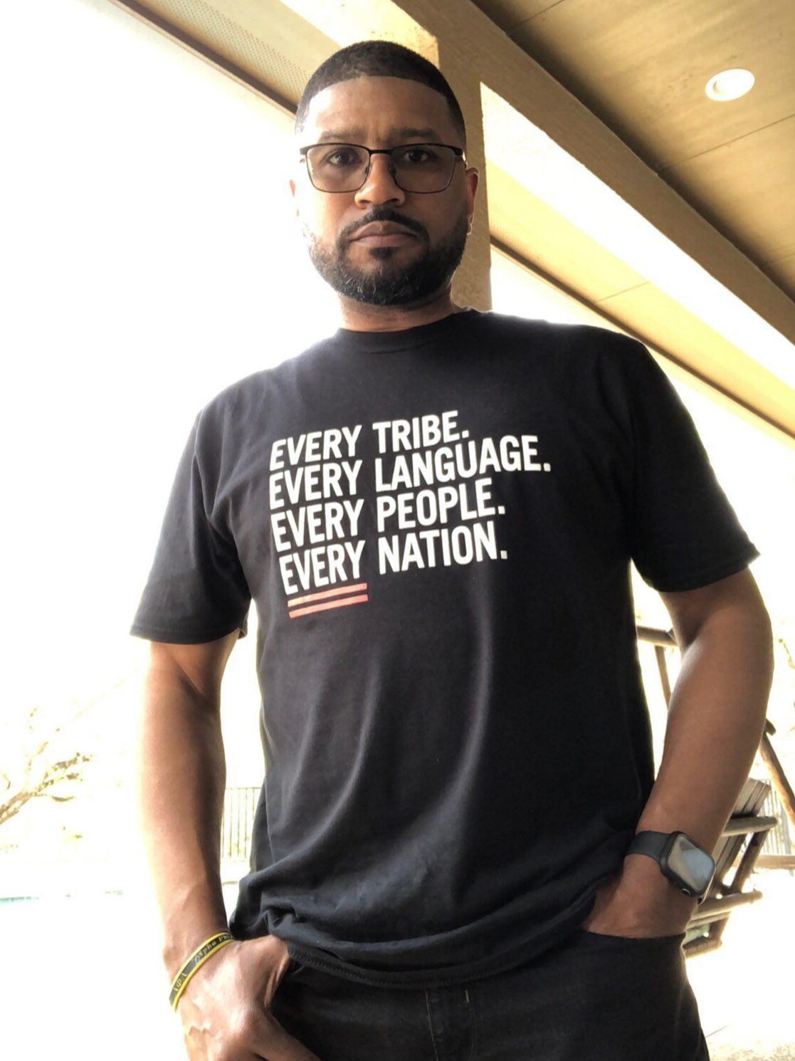 Man wearing glasses and a black "every tribe. every language. every people. every nation." t-shirt with text advocating for inclusivity.
