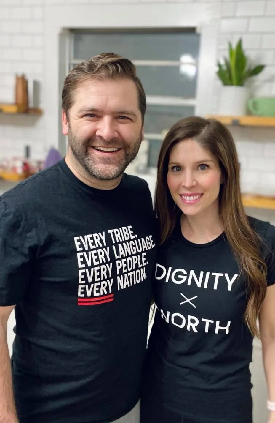 Two people smiling wearing black t-shirts with text. The man's t-shirt reads "Every tribe. Every language. Every people. Every nation." The woman’s t-shirt reads "Dignity x North."