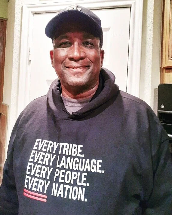 Smiling man wearing a navy blue hoodie with text: 'Every tribe. Every language. Every people. Every nation.' and a baseball cap.