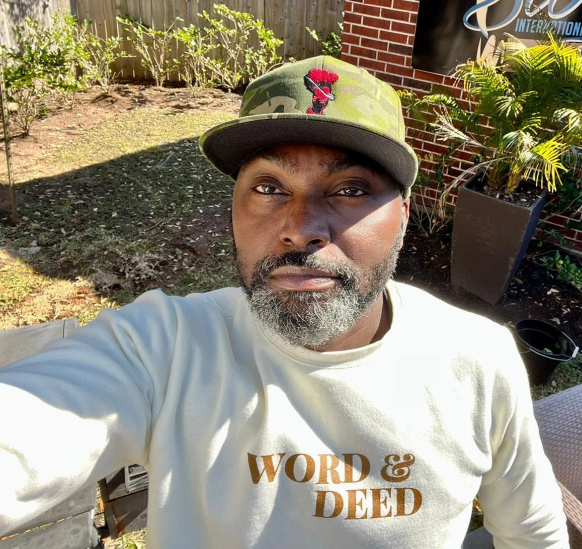Man wearing a camo cap and a white shirt with 'Word & Deed' text, standing outdoors with plants and a brick wall in the background.