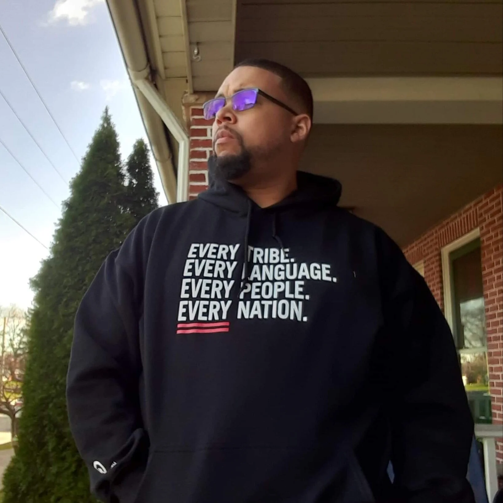 Person in black hoodie with text: 'Every tribe. Every language. Every people. Every nation.' standing outdoors near a brick wall and a tree, looking away.