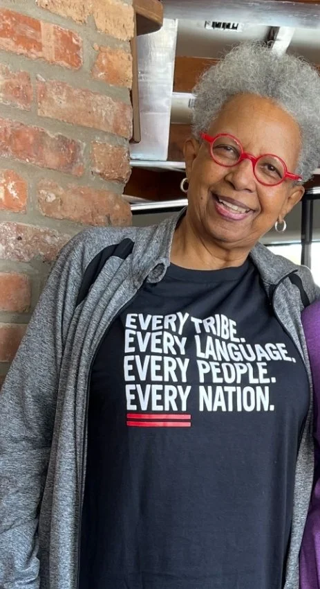Smiling person wearing red glasses and a black T-shirt with the message "Every tribe. Every language. Every people. Every nation."