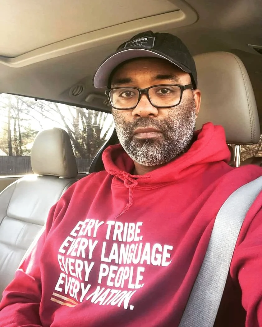 Man wearing glasses and a red "Every Tribe. Every Language. Every People. Every Nation." hoodie with text in a car.