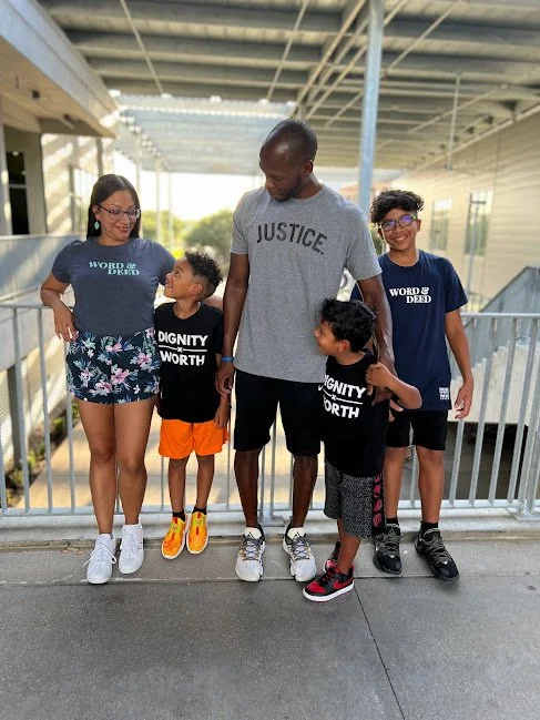 A family of five standing together on a pedestrian bridge. The mother is wearing a floral skirt with a "Word and Deed" T-shirt. The father is wearing athletic shorts and a "Justice" T-shirt. Their three children, two boys and a smaller child, are wea