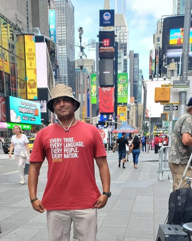 Man in red T-shirt with "Every Tribe. Every Language. Every People. Every Nation." text, wearing a hat, standing in Times Square, New York City. Background includes buildings and colorful billboards.