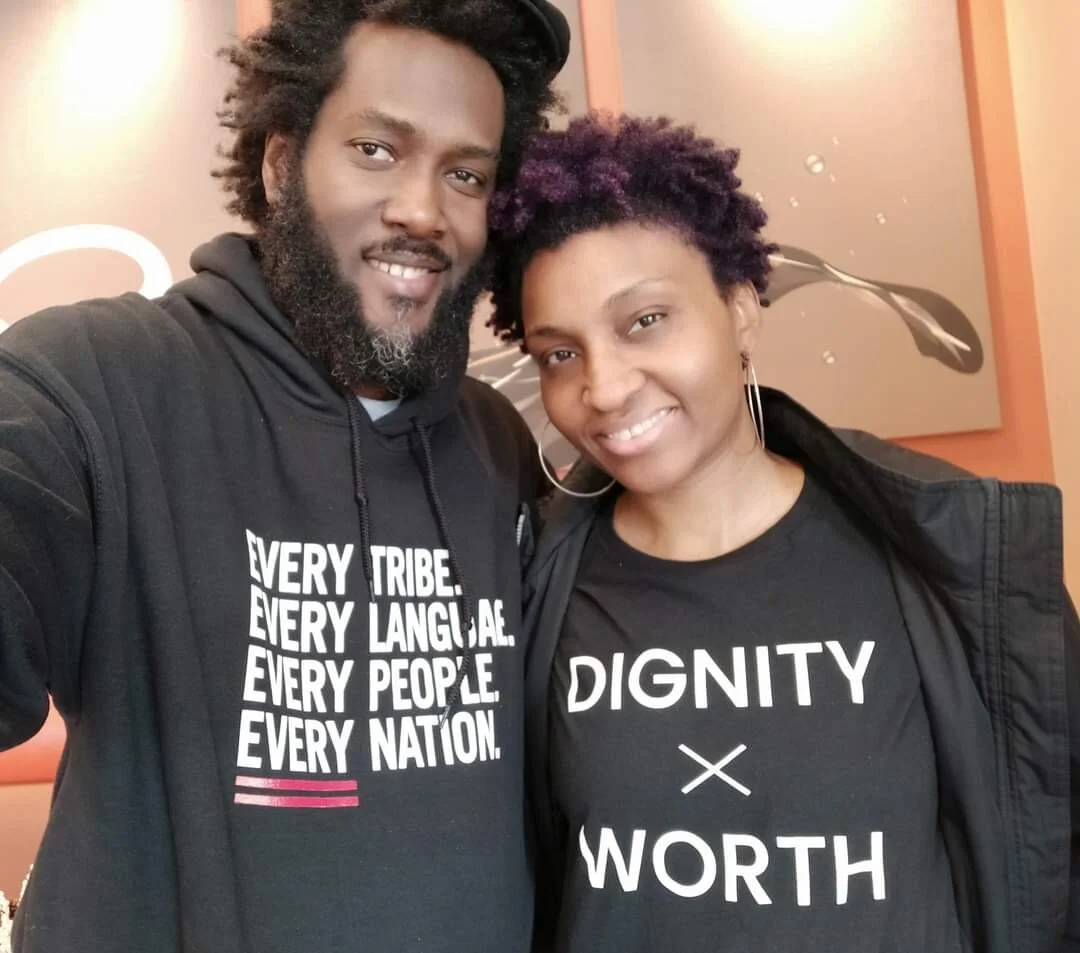 A man and a woman smiling, wearing black tops with text. The man's shirt says "Every tribe. Every language. Every people. Every nation." The woman's shirt says "Dignity x Worth."