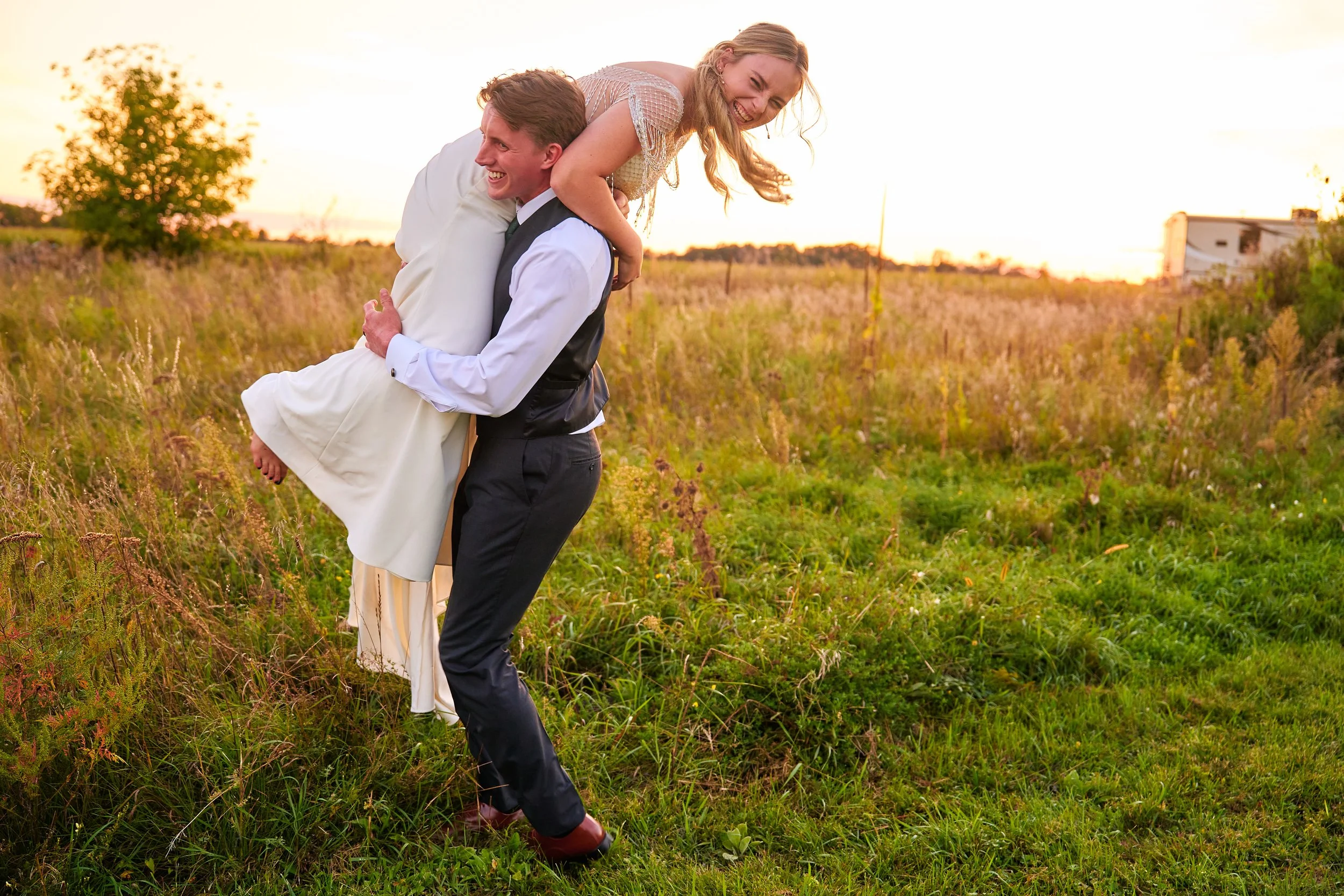 A couple is outdoors at sunset, with the man carrying the woman on his shoulders in a grassy field, both smiling and laughing.