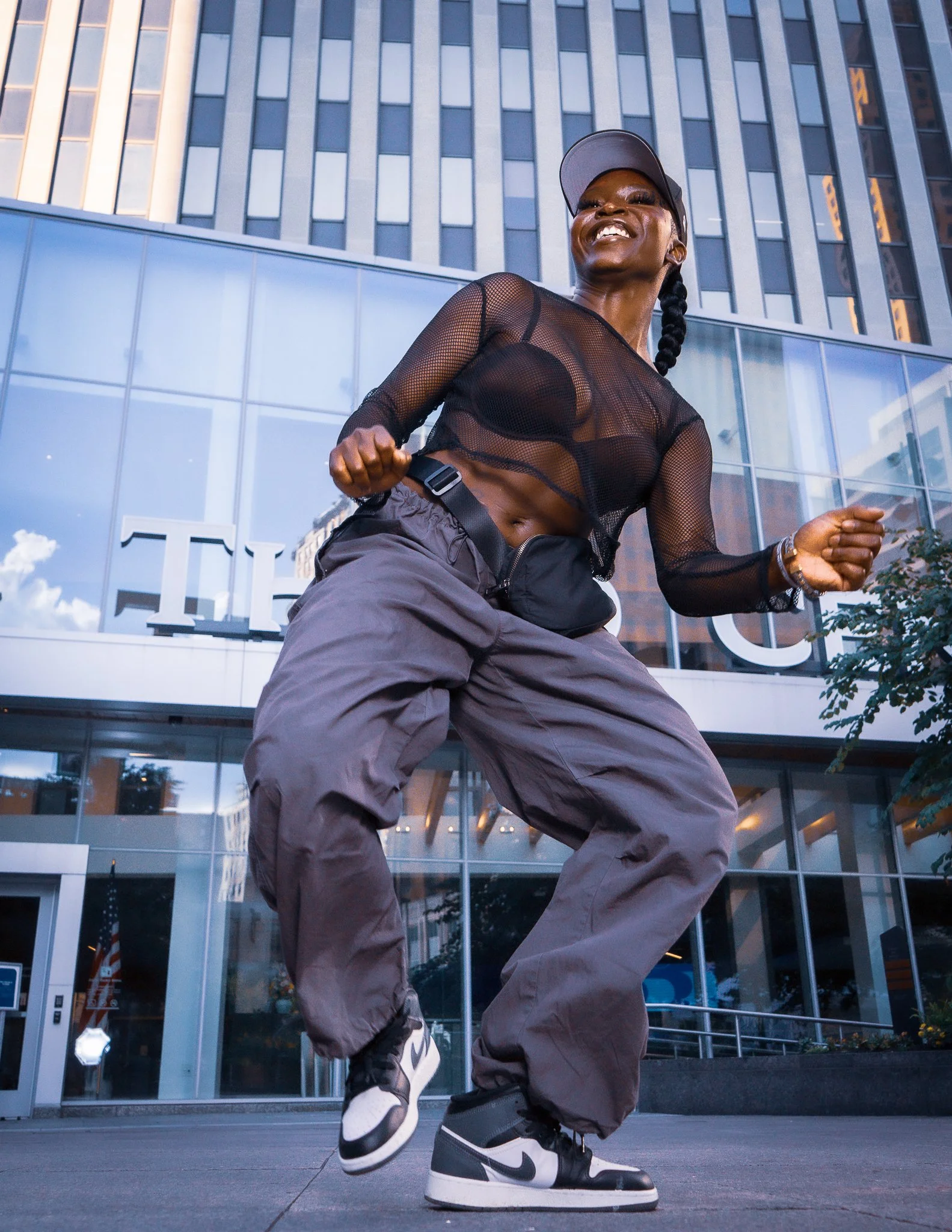 A woman dancing outdoors in front of a modern glass building, wearing a black mesh top, gray baggy pants, and black and white sneakers, smiling happily.