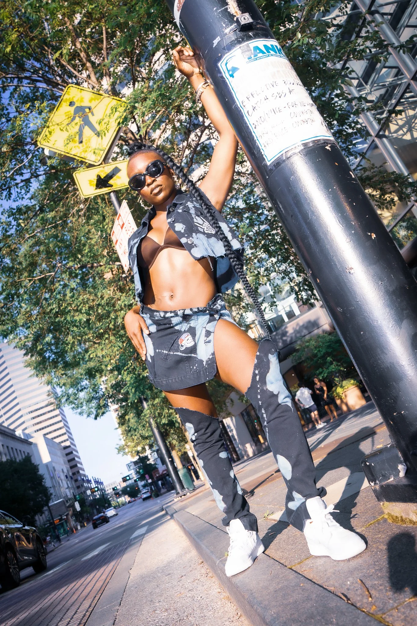 A woman in black and white tie-dye clothing, sunglasses, and white sneakers stands on a city sidewalk, leaning against a black pole, with trees and tall buildings in the background.