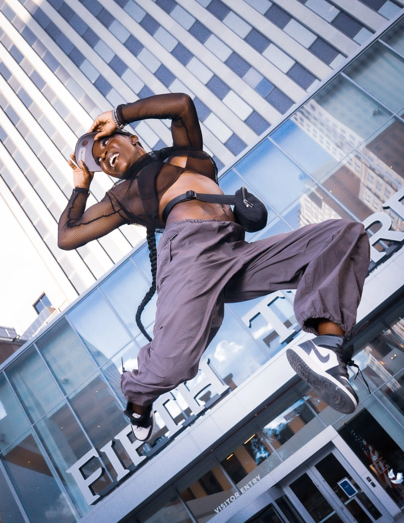 A person jumping in front of a modern building with a glass facade, wearing a sheer black top, gray loose pants, black sneakers, and a small black waist bag, smiling and holding a gray cap.
