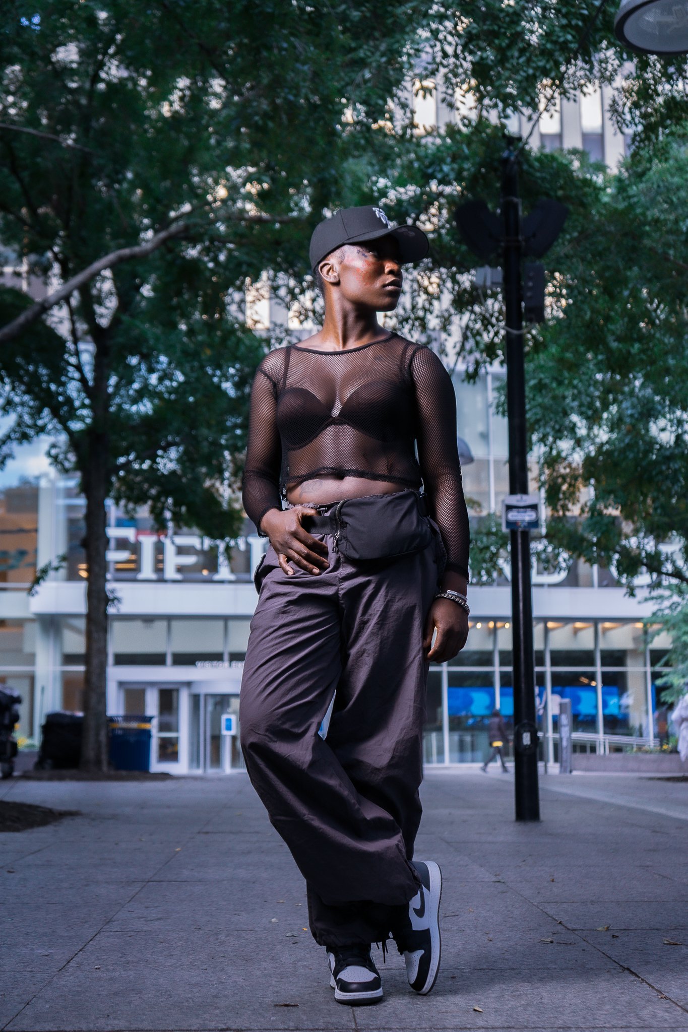A person standing outdoors on a city sidewalk, wearing a black baseball cap, a black mesh long-sleeve top over a black bra, baggy black pants, and black and white Nike sneakers. The background shows trees, a building with glass windows, and a street 