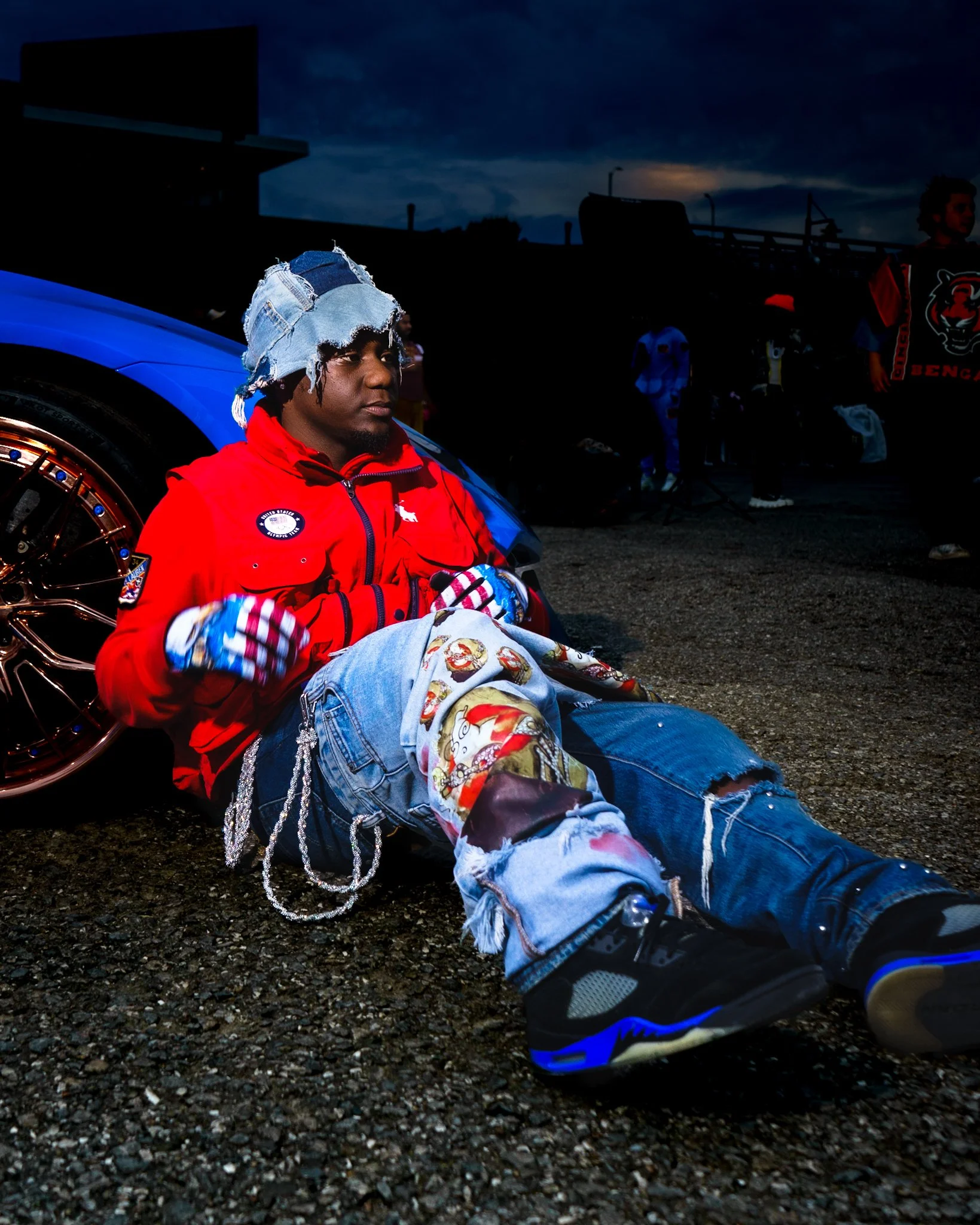 Young man sitting on the ground next to a blue car at dusk, wearing a red jacket, patched jeans, and a denim hat.
