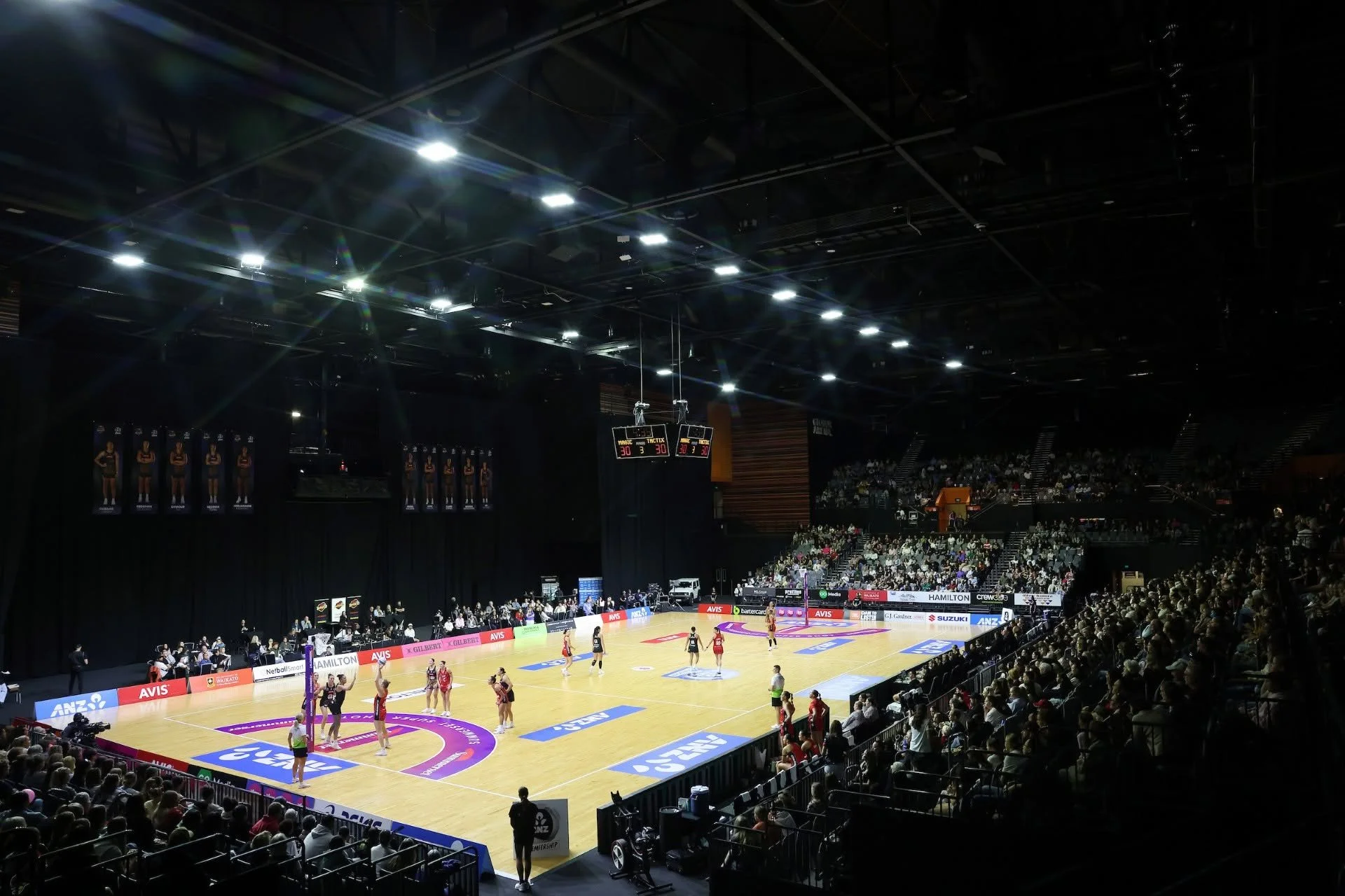 An indoor basketball game with players on the court, audience in the stands, and bright arena lighting.