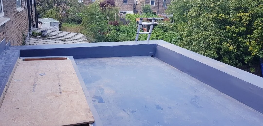 A rooftop in Newport Pagnell with a flat Lead grey single ply roof surface, some plywood, and a ladder leading to the edge, surrounded by trees and neighboring buildings.