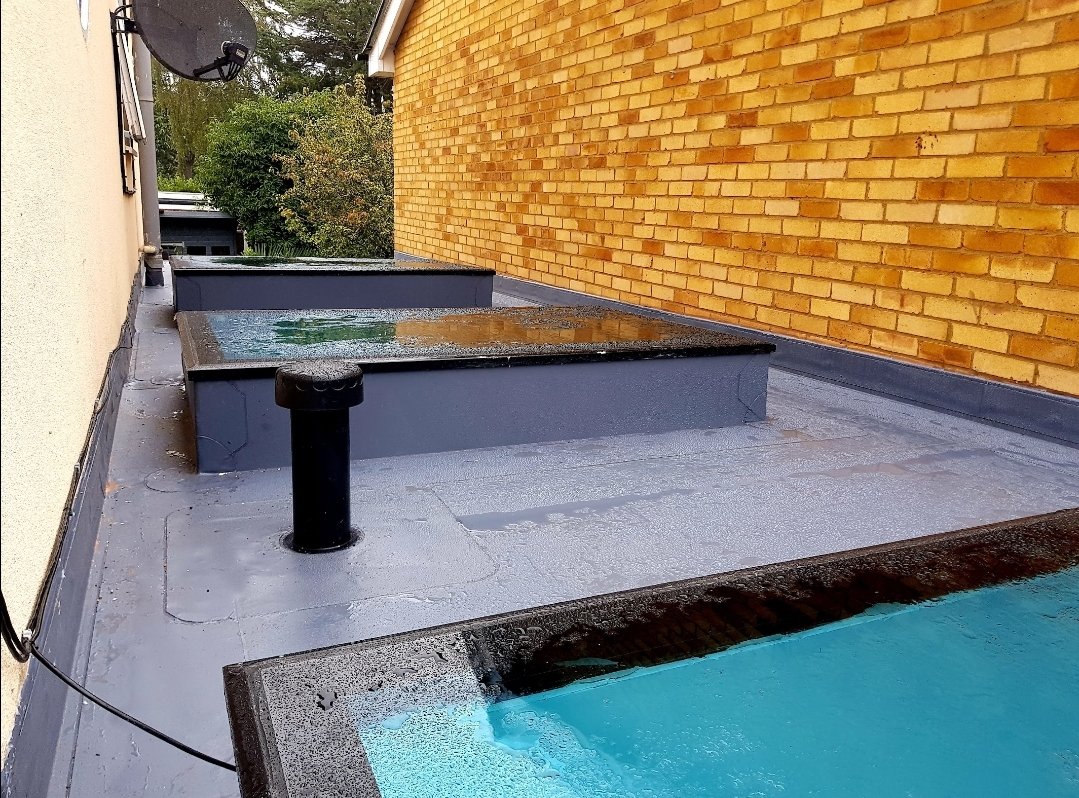 View of a rooftop with a small pool, some water on a raised platform, a black vent pipe, and a satellite dish, surrounded by brick and siding walls with trees in the background.