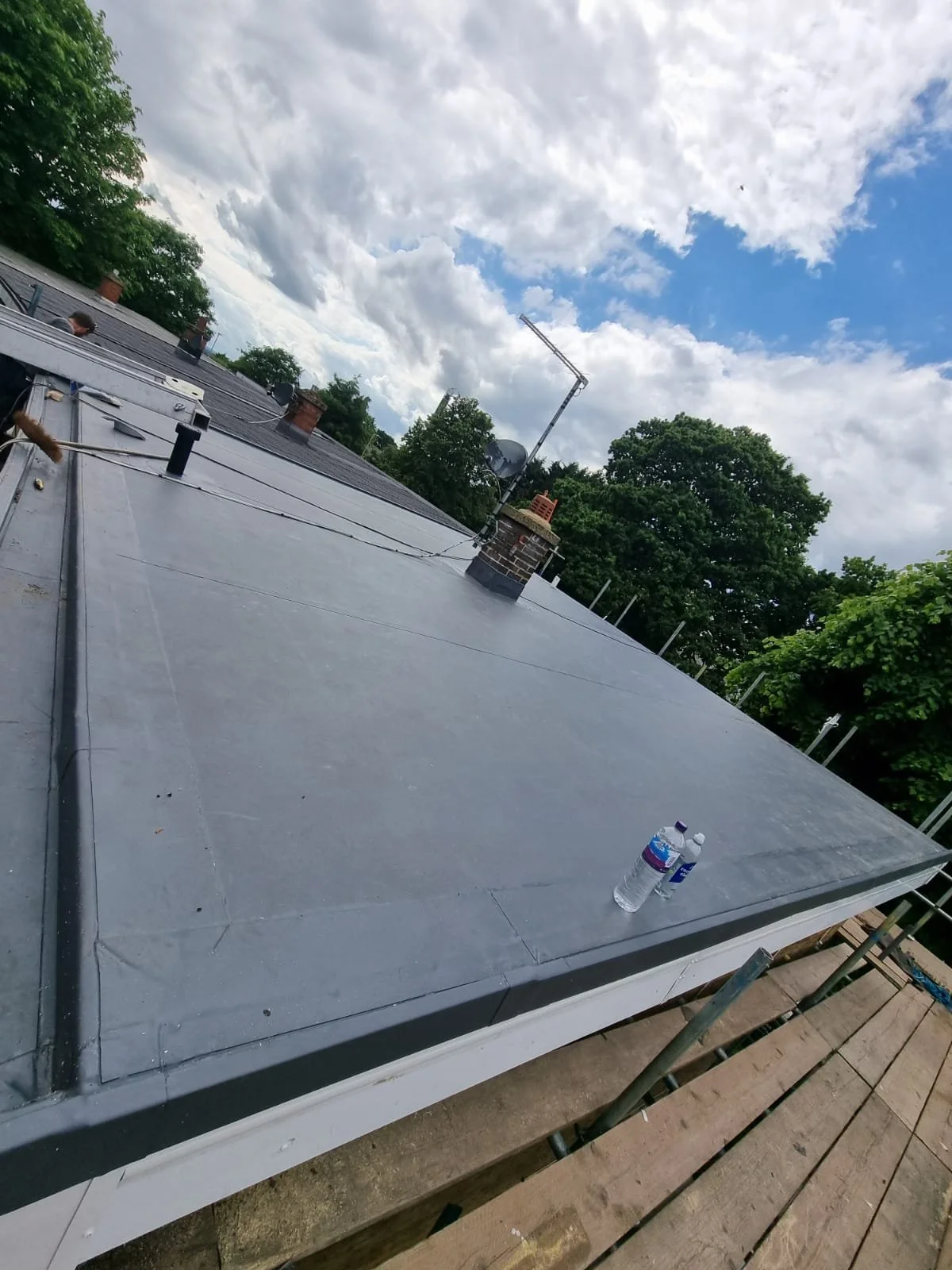 Photo of a flat rooftop with a water bottle and a plastic container near the edge. The sky is partly cloudy with some blue visible, and there are trees and chimneys on surrounding rooftops.