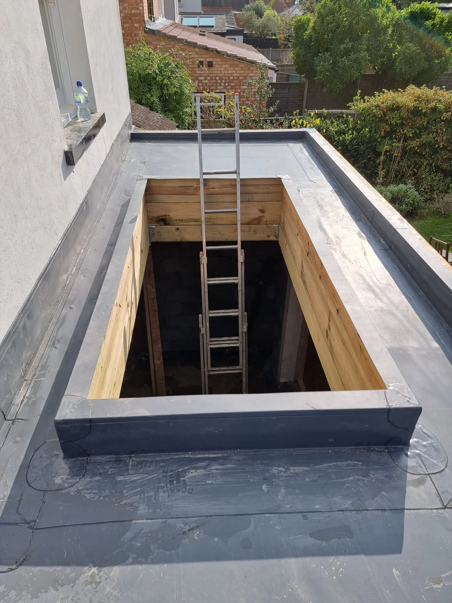 Construction site with a square opening in the roof, surrounded by a metallic frame, and a ladder leading down into the opening. The area is being prepared for roof work or installation.