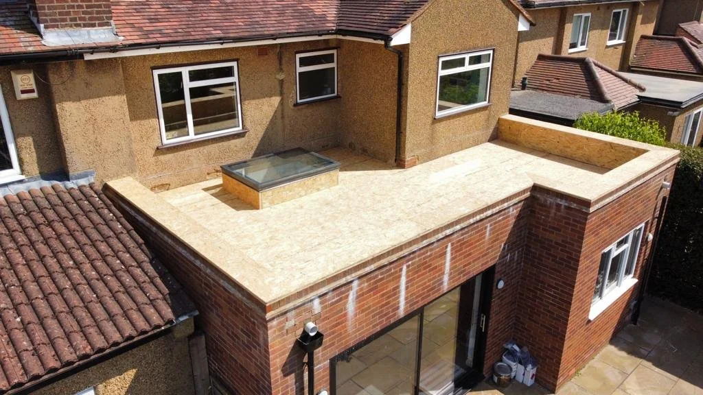 A rooftop terrace covered in 18mm ply in Milton Keynes enclosed by a brick wall, and a small skylight window. The terrace is attached to a multi-story brick residential building with several windows.