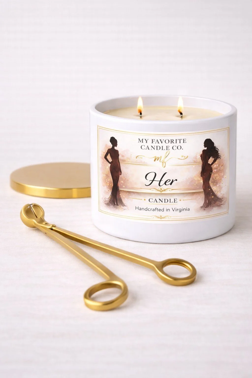 A white candle with two flames in a decorative label featuring silhouettes of women, labeled "My Favorite Candle Co. Her," with a gold soap bar, a gold wax spatula, and a white background.