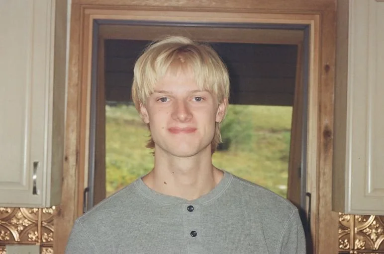 A young man standing inside a room near a window with a wooden frame, showing a green outdoor landscape in the background.