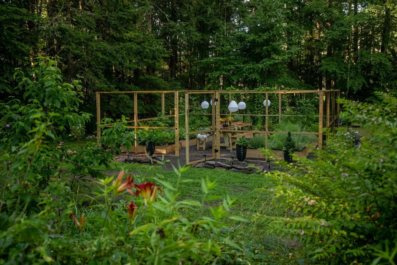 A backyard garden enclosed by wooden fencing with plants and flowers, outdoor table with sunflower decoration, and hanging lanterns, surrounded by trees and greenery.
