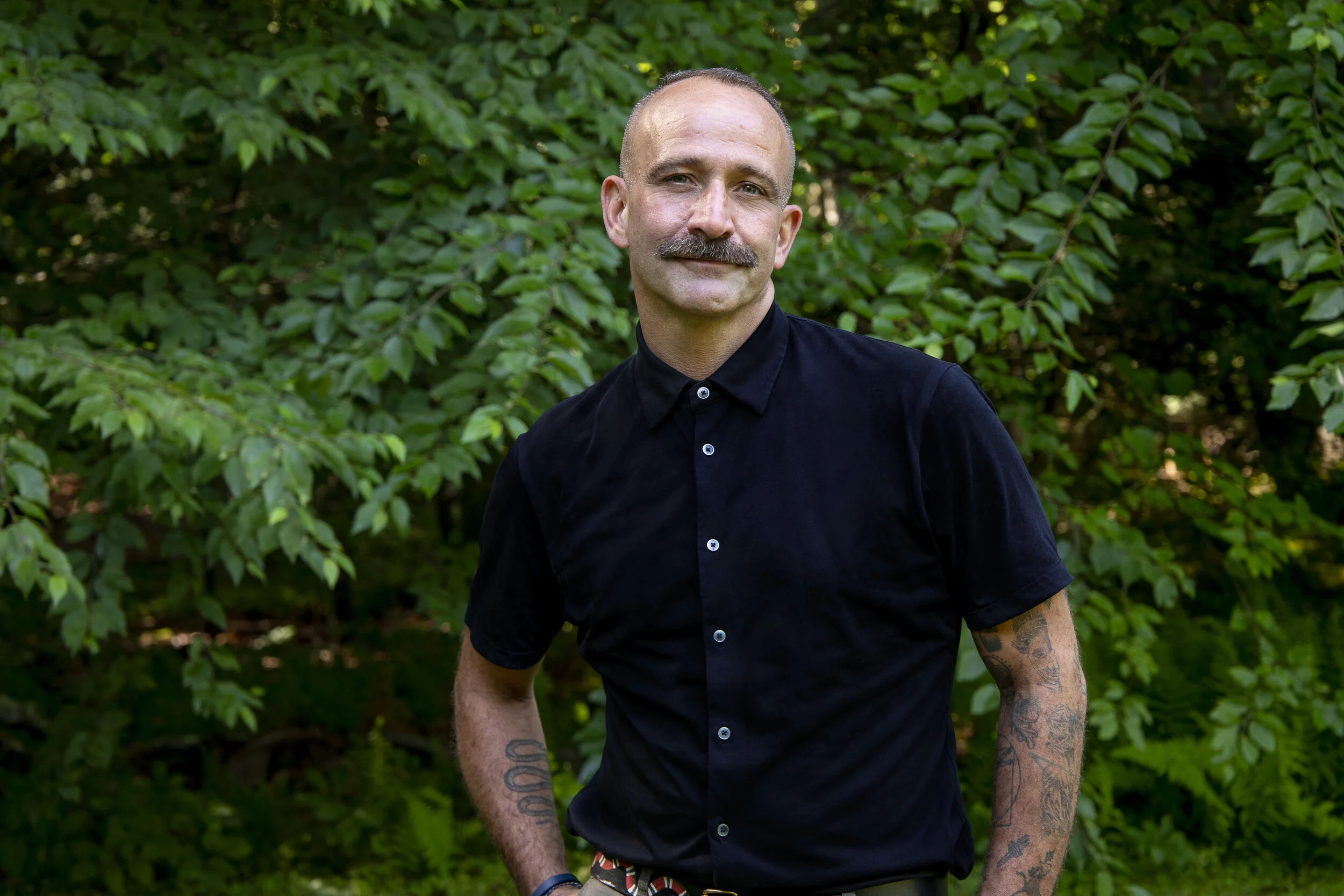 A man with a mustache and tattoos on his arms, wearing a black short-sleeved button-up shirt, standing outdoors in front of green foliage.