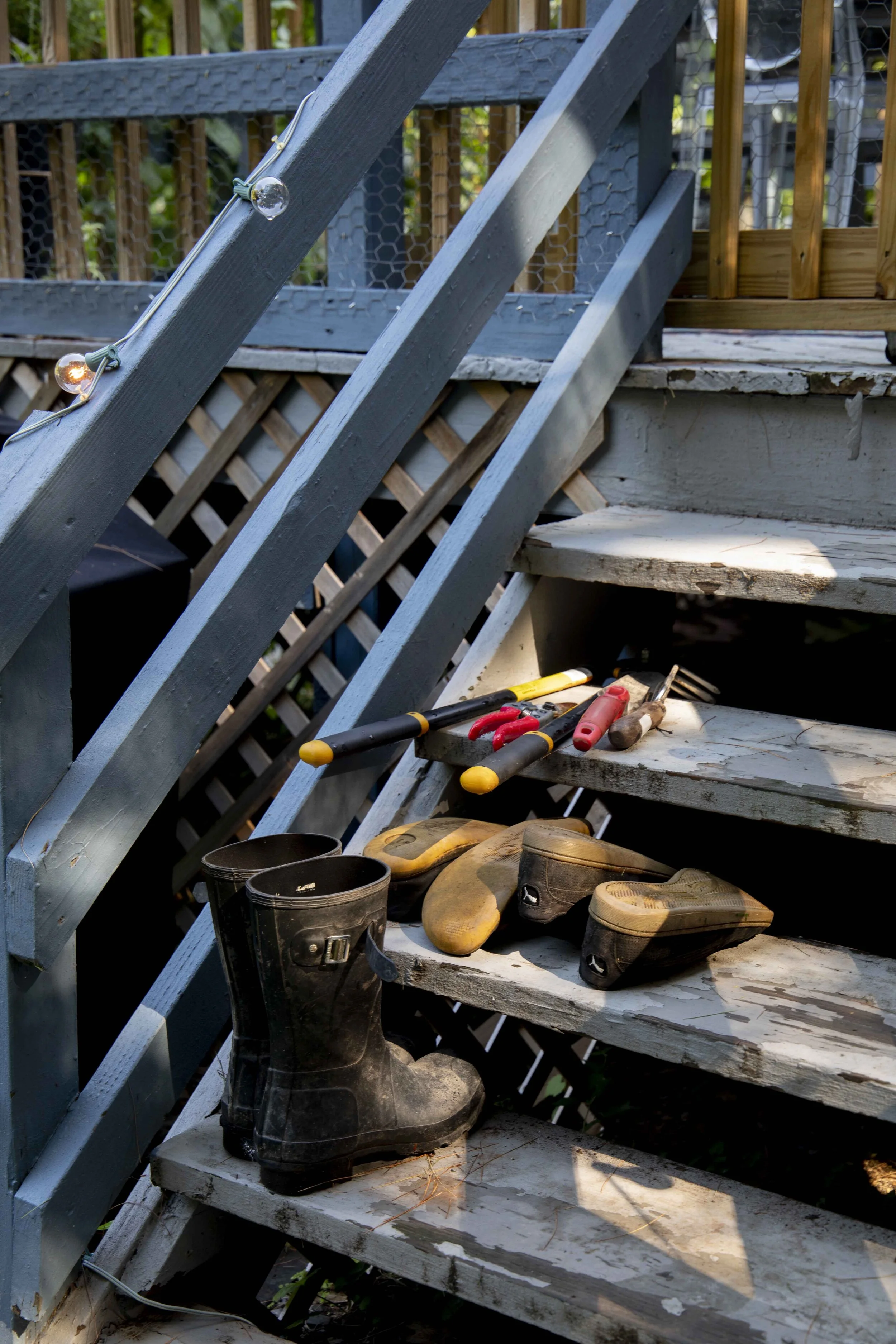Outdoor wooden staircase with gardening tools, including a pair of rubber boots, two gardening shovels, and hedge shears, placed on the steps.