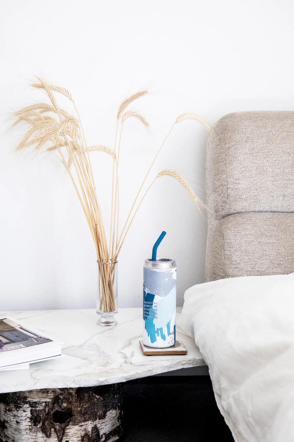 A bedside table with a glass vase holding dried wheat stalks, a magazine, a decorated reusable water bottle with a blue straw on a coaster, a part of a bed with white bedding, and a beige upholstered headboard against a white wall.