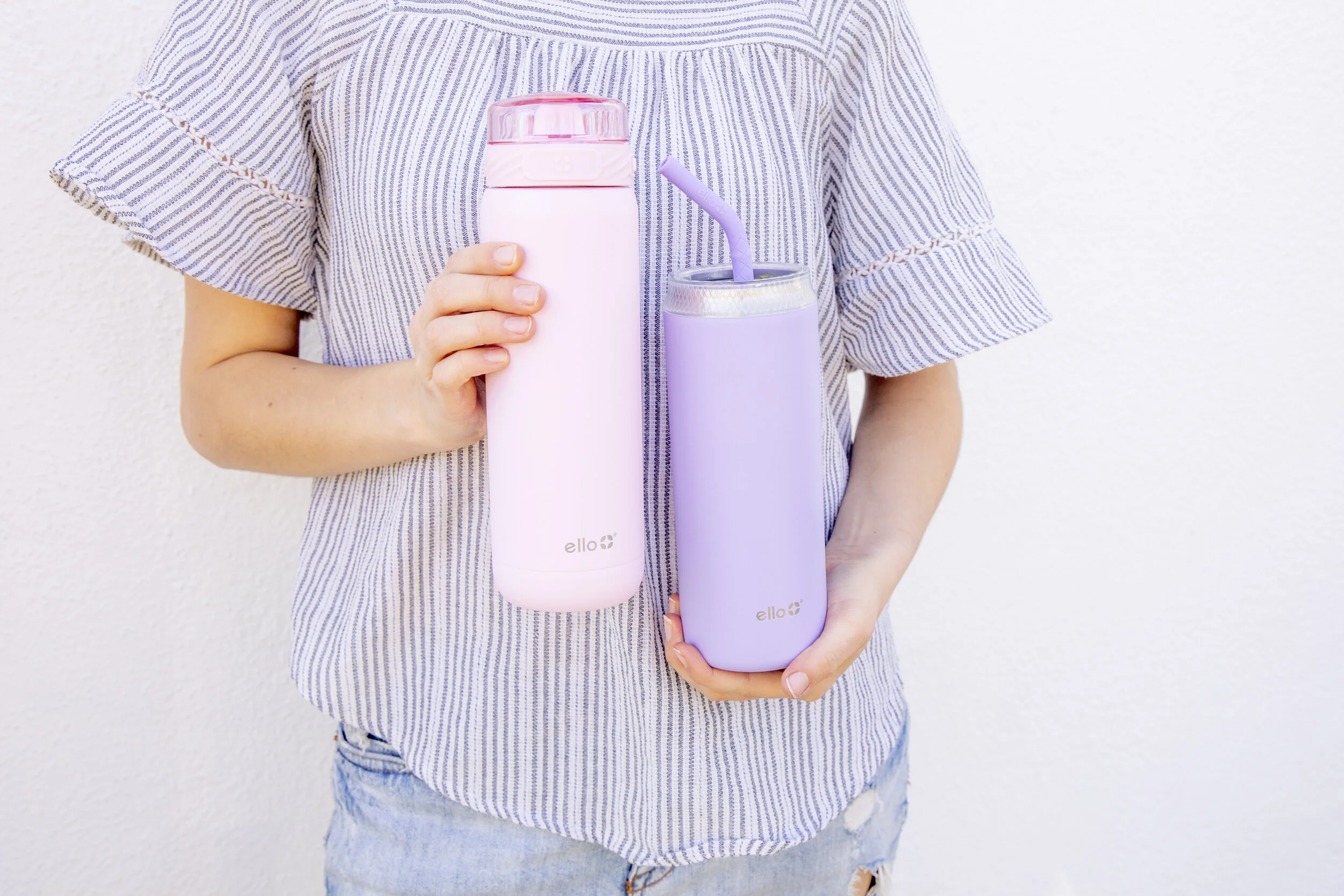 Person holding two pastel-colored reusable bottles, one pink and one purple, against a white wall.