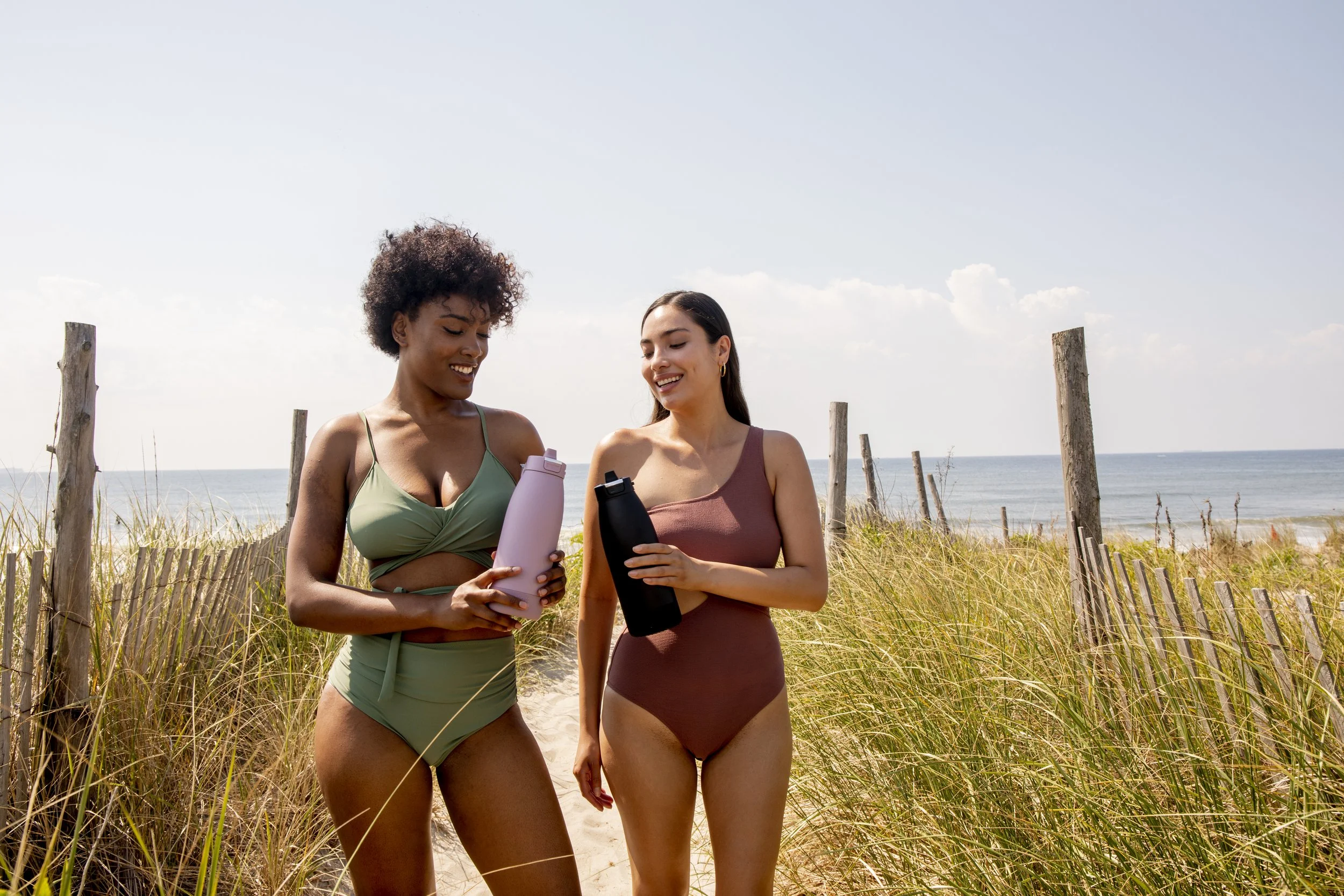 Two women in swimsuits walking on the beach with a wooden fence and ocean in the background, holding water bottles and smiling.