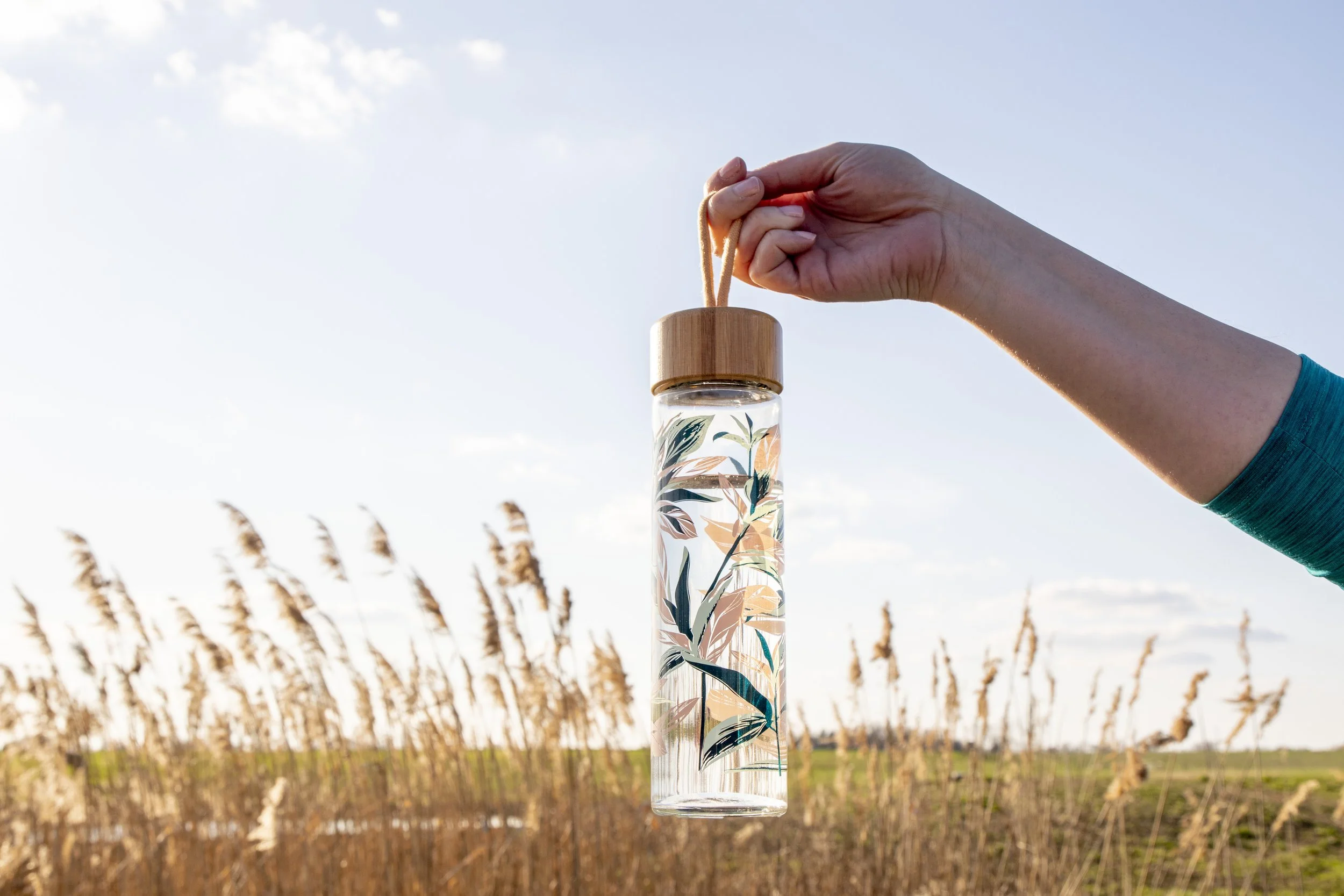 A hand holding a glass water bottle with a wooden cap and a strap, featuring a botanical pattern, against a background of a field with tall grass and a blue sky.