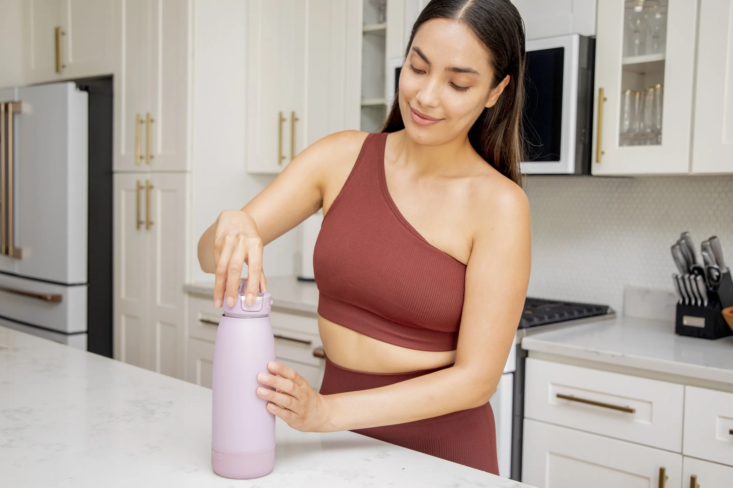 A woman in a rust-colored sports bra and matching high-waisted leggings opening a pink reusable water bottle in a modern kitchen.