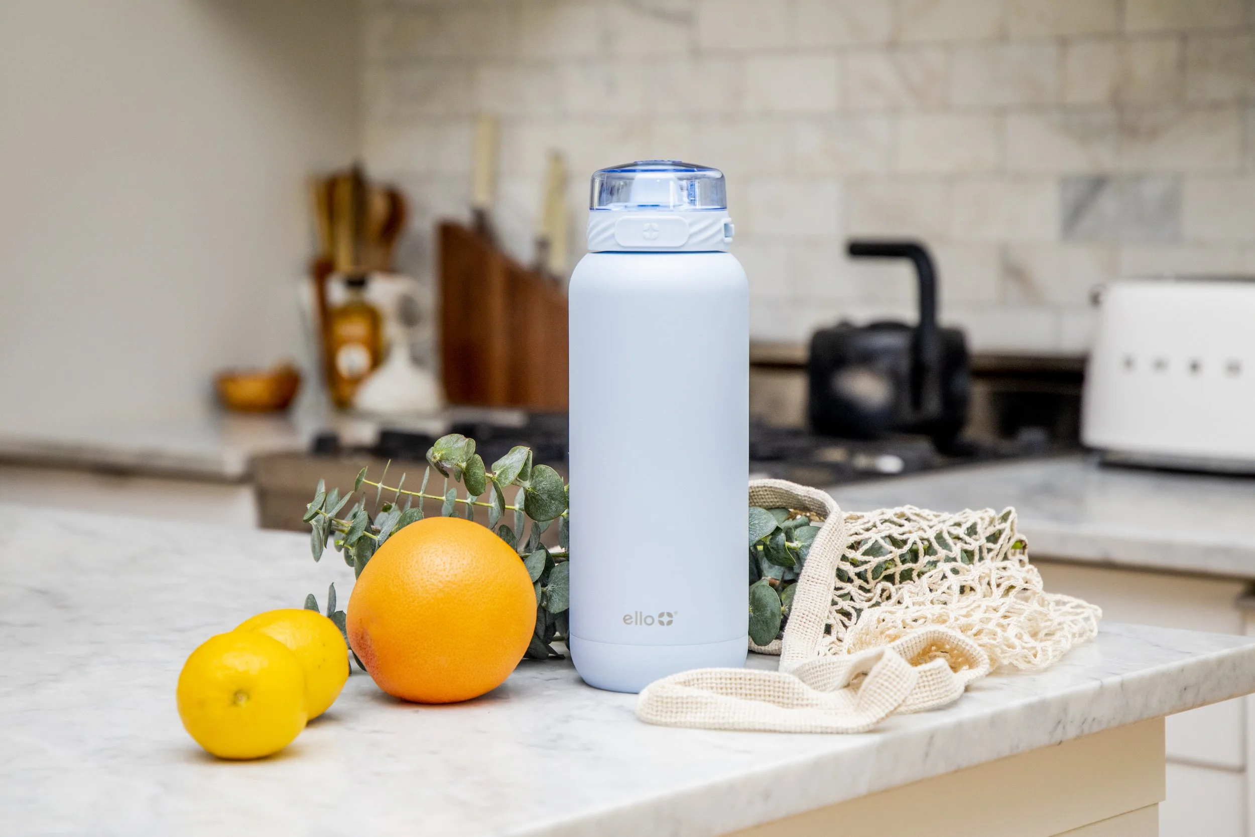 A white insulated water bottle on a kitchen countertop with an orange, a yellow lemon, green leaves, a reusable mesh bag, and a beige cloth, with a stove, kettle, and kitchen utensils in the background.
