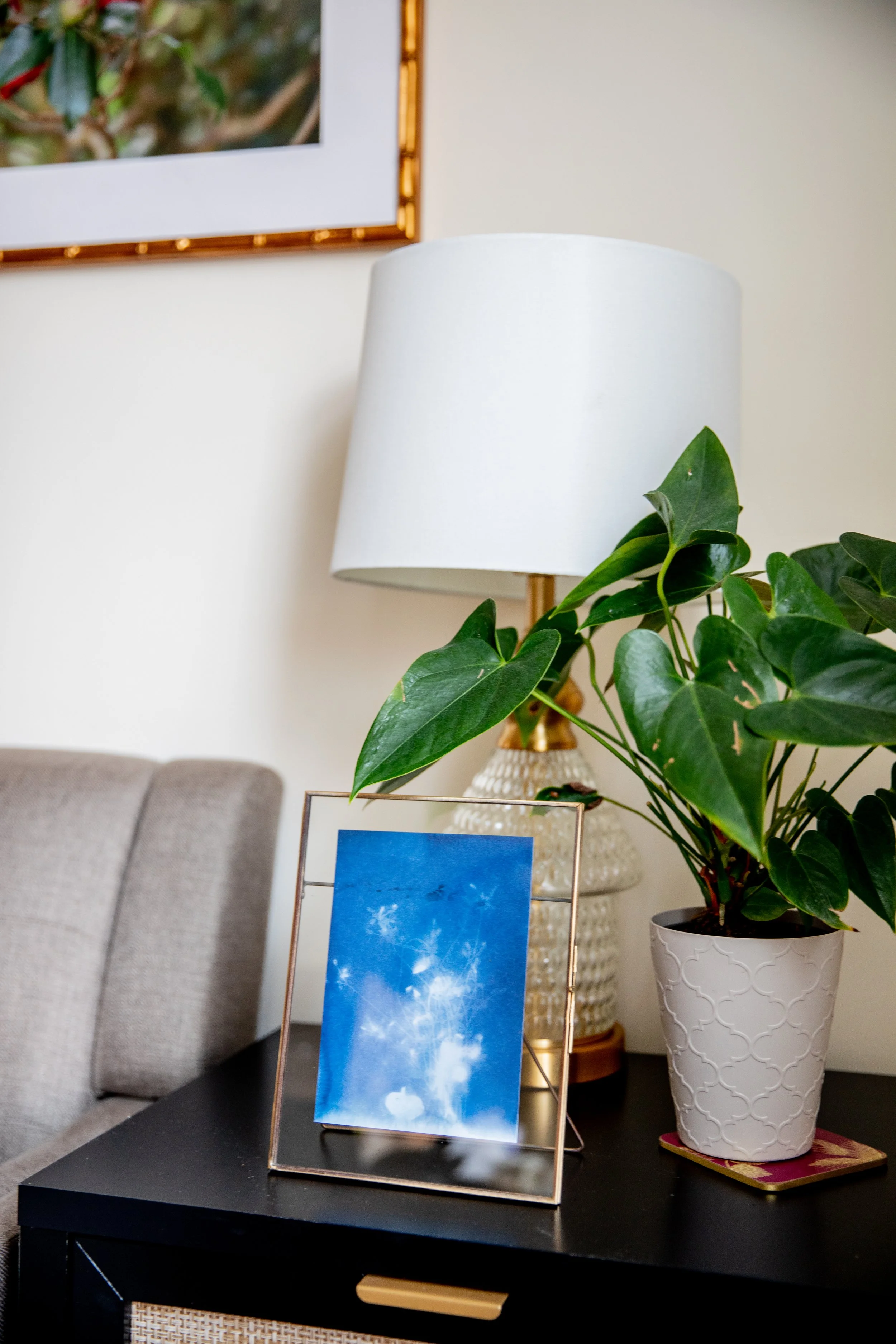 A living room side table holding a potted plant with large green leaves, a table lamp with a white shade, a framed photo with a blue sky and white clouds, and a framed artwork hanging on the wall above.