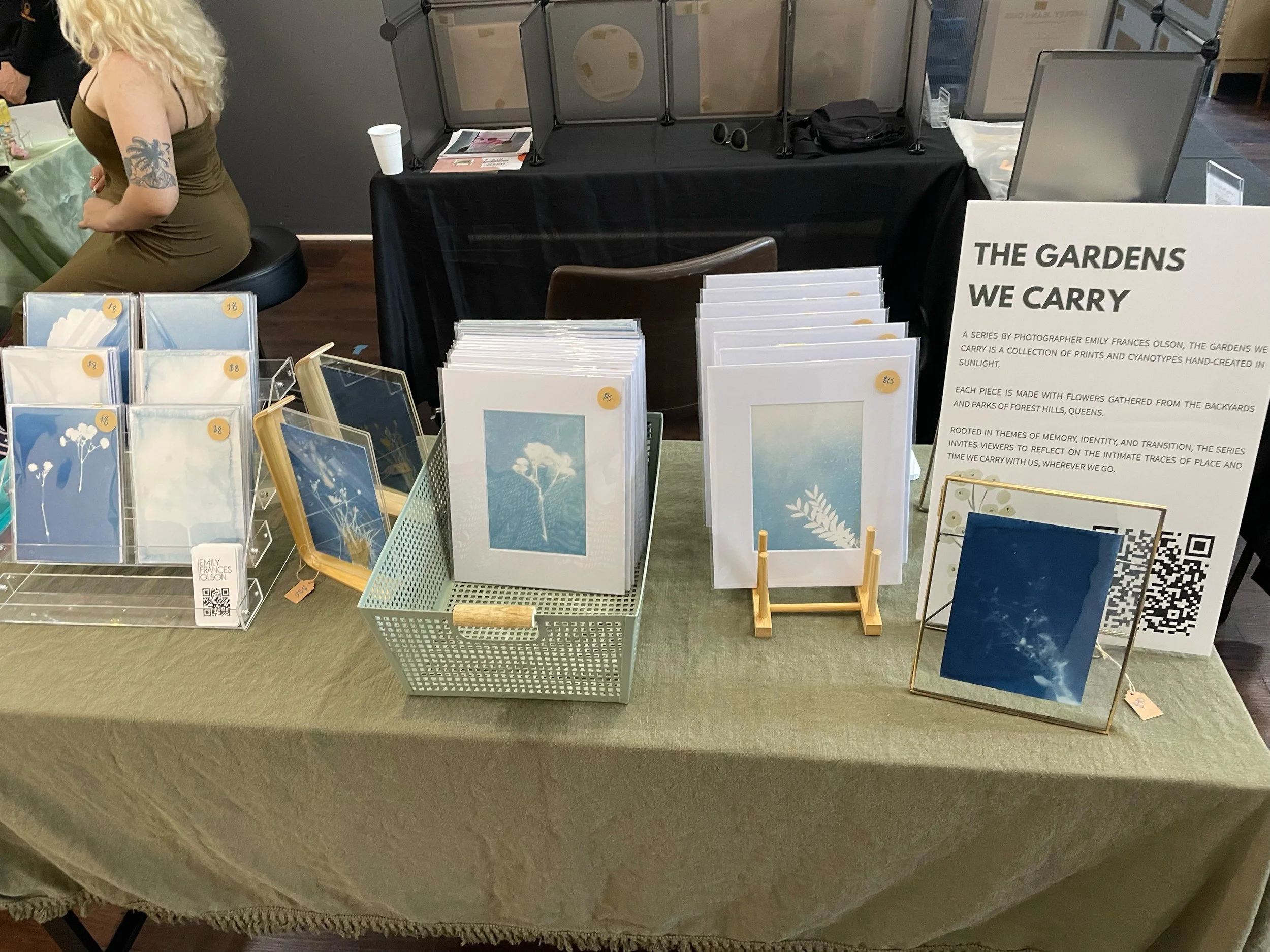 Table display of prints and cyanotypes titled 'The Gardens We Carry' by photographer Emily Frances Olson. The display includes various prints in frames and pouches, a sign explaining the series, and a QR code for more information.