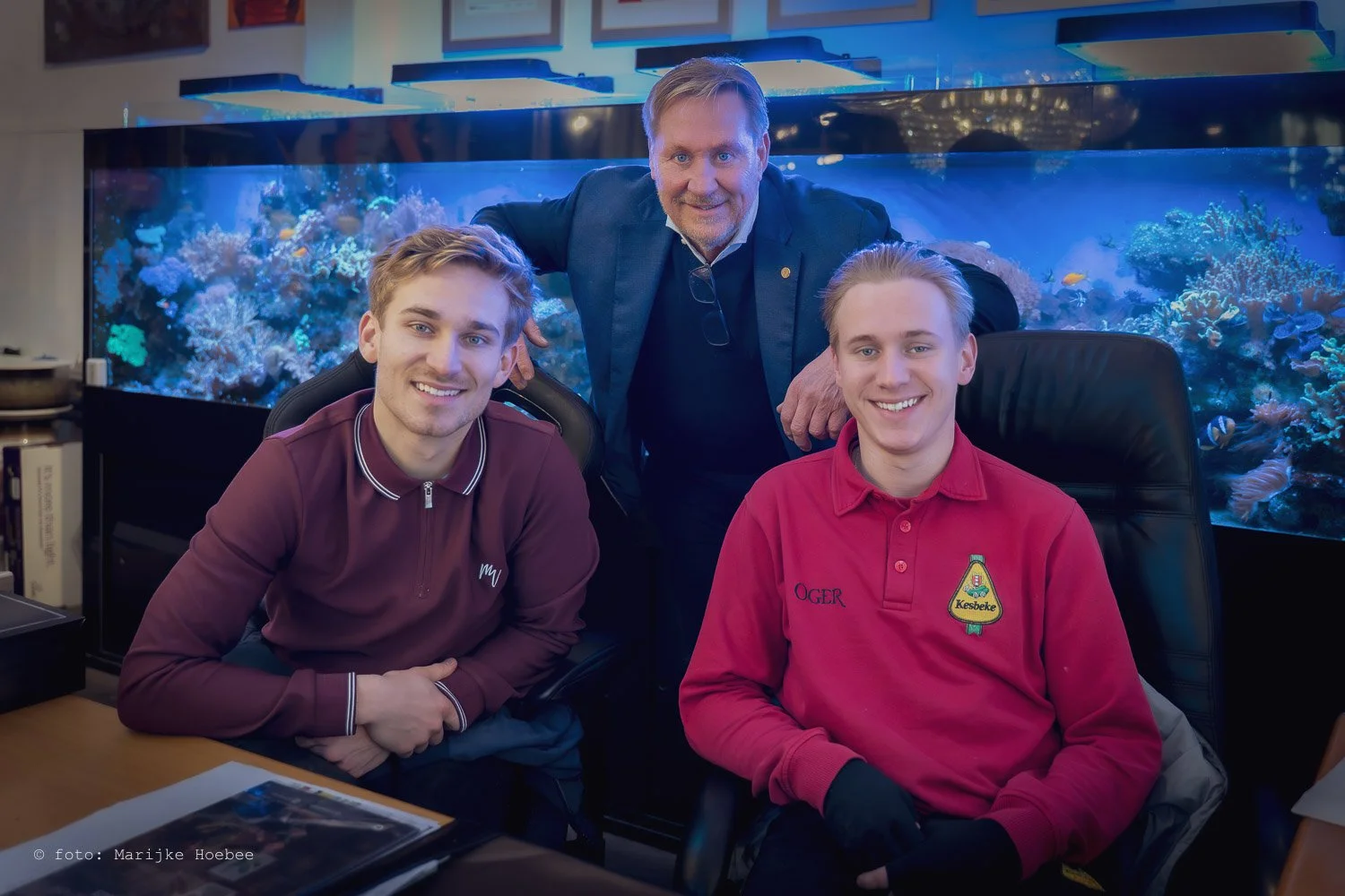 Drie mannen in een kantoor met een groot aquarium op de achtergrond, twee mannen zitten en één staat achter hen, lachend.