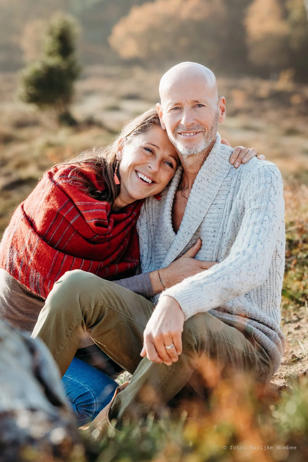 Een gelukkige oude man en vrouw zitten buiten op een herfstlandschap, de vrouw knuffelt de man en beiden glimlachen.