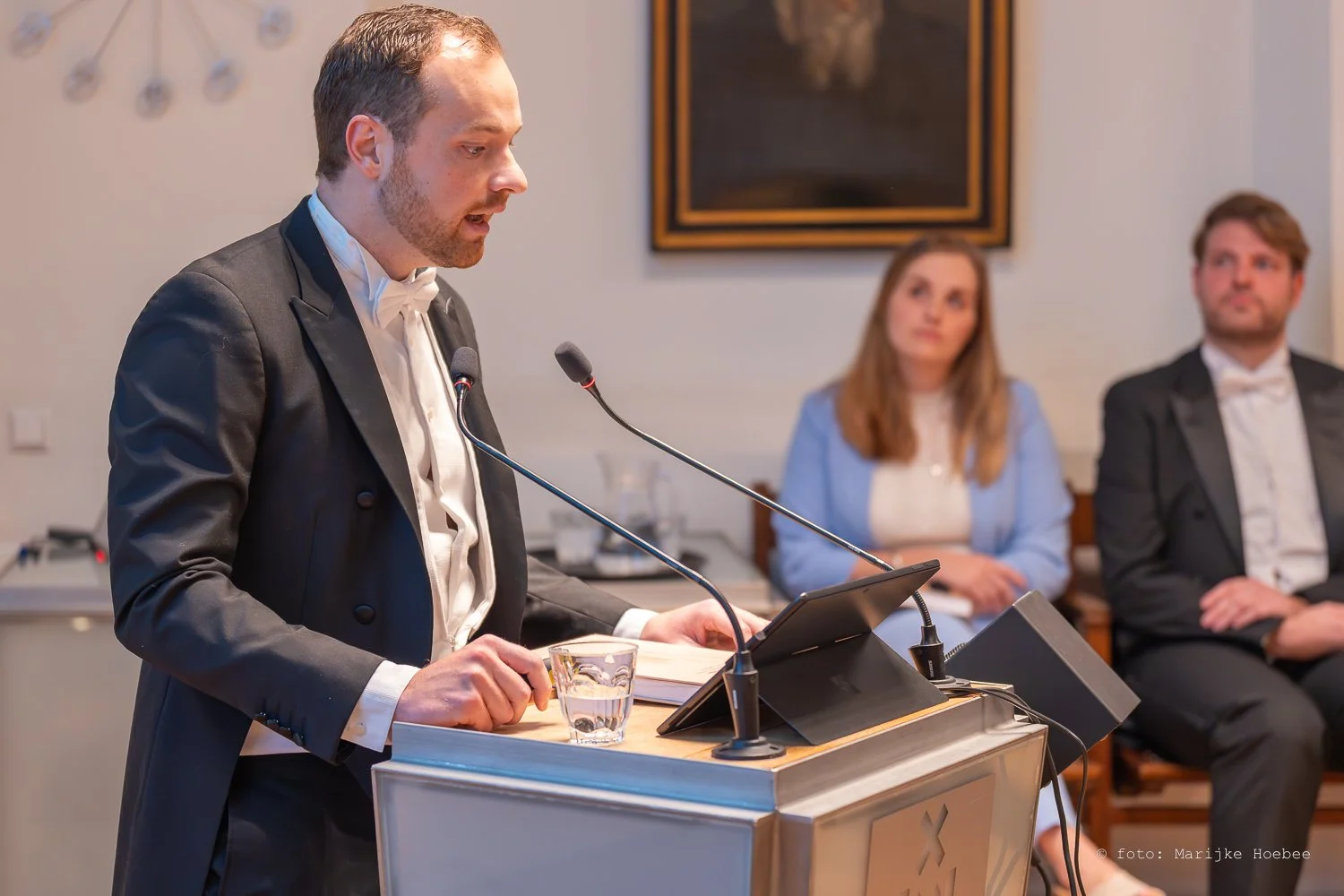 Man in een tuxedo die voor een microfoon spreekt achter een lessenaar, met twee luisteraars op de achtergrond in een formele setting.