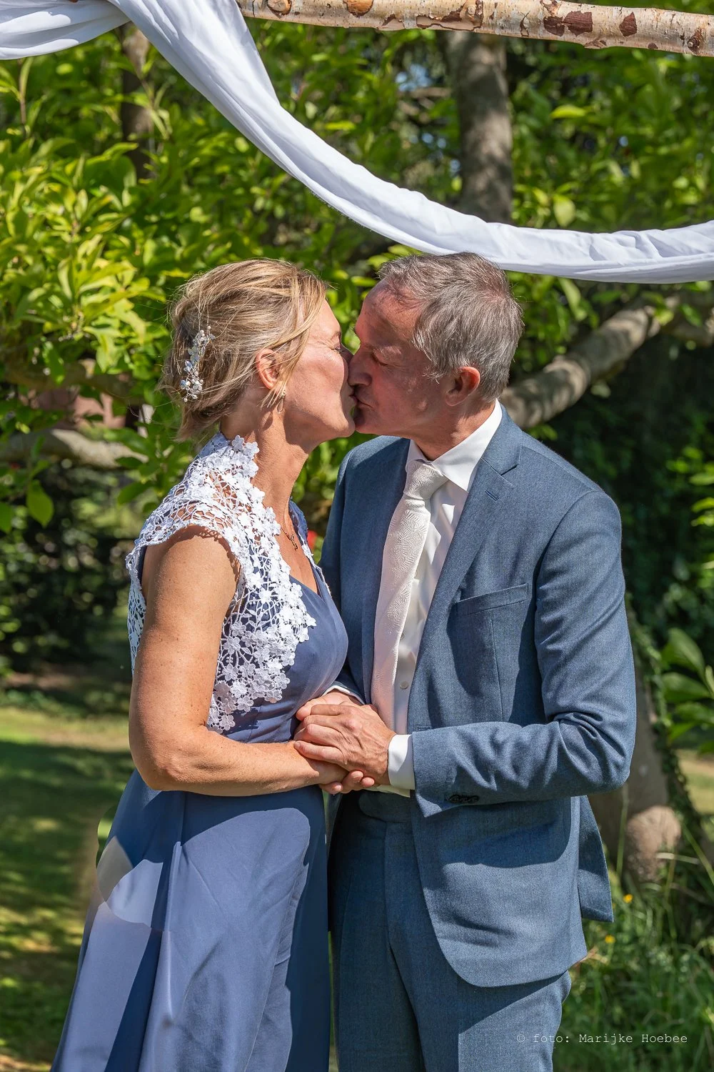 Een omhelzend echtpaar dat elkaar kust tijdens een buitenhuwelijk, omringd door groene bomen en zonlicht.
