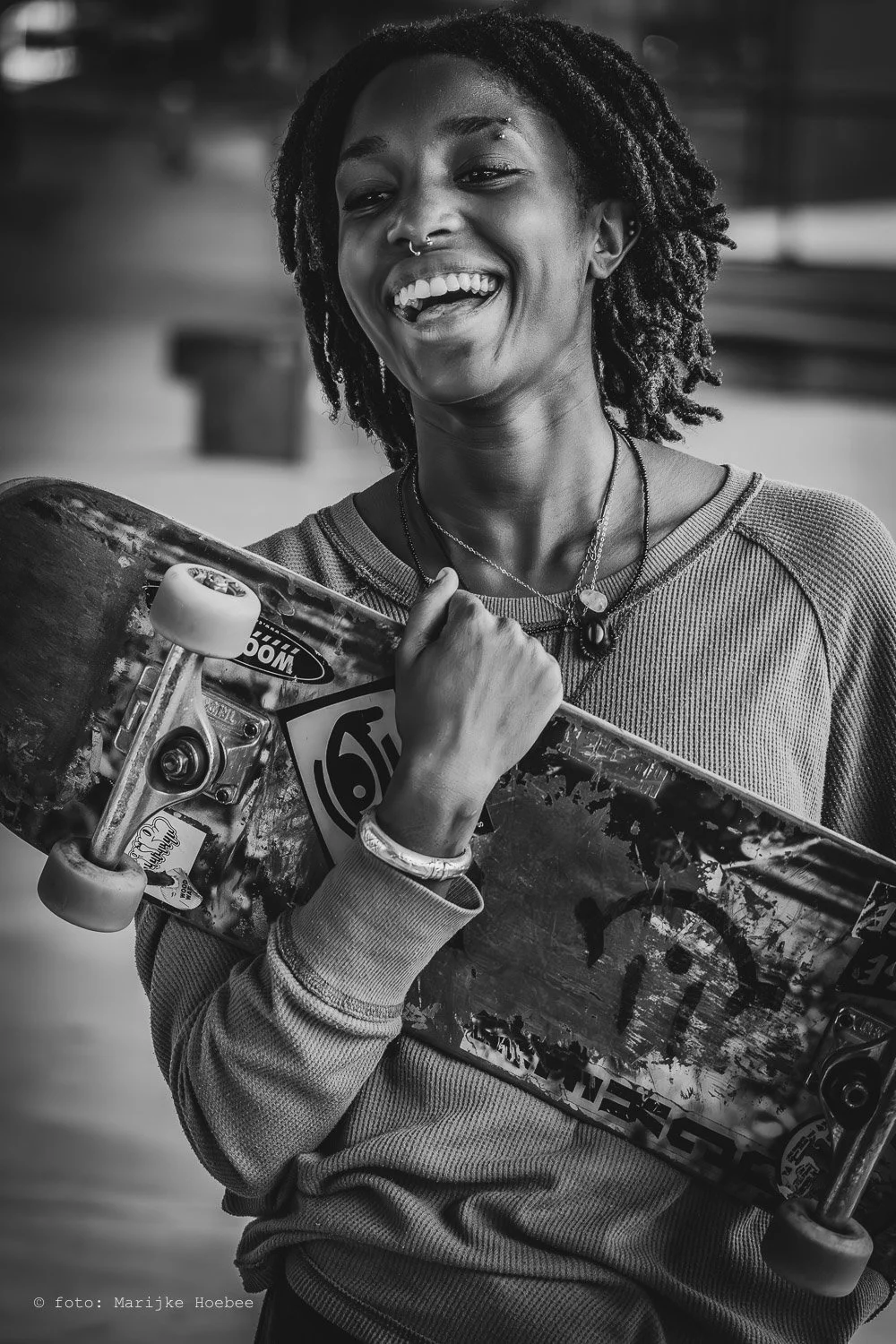 Een lachende vrouw met dreadlocks houdt een skateboard onder haar arm.