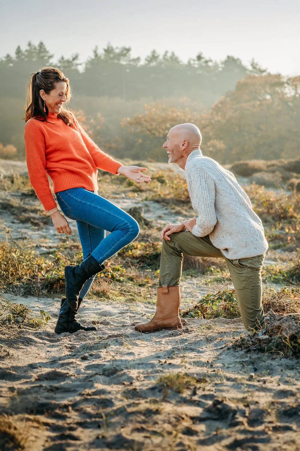 Een vrouw en een man lachen samen in een bos, de vrouw staat op één been en de man knielt voor haar, beiden in sweaters en laarzen, tijdens zonsondergang.