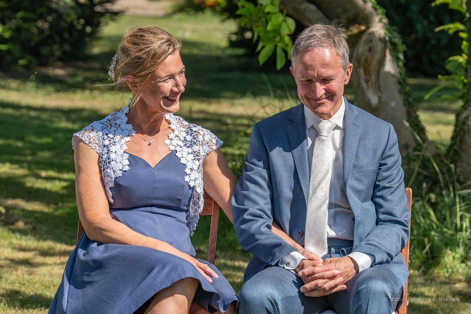 Een oudere vrouw en een oudere man zitten samen in een tuin, lachend en gekleed voor een speciale gelegenheid, mogelijk een bruiloft.
