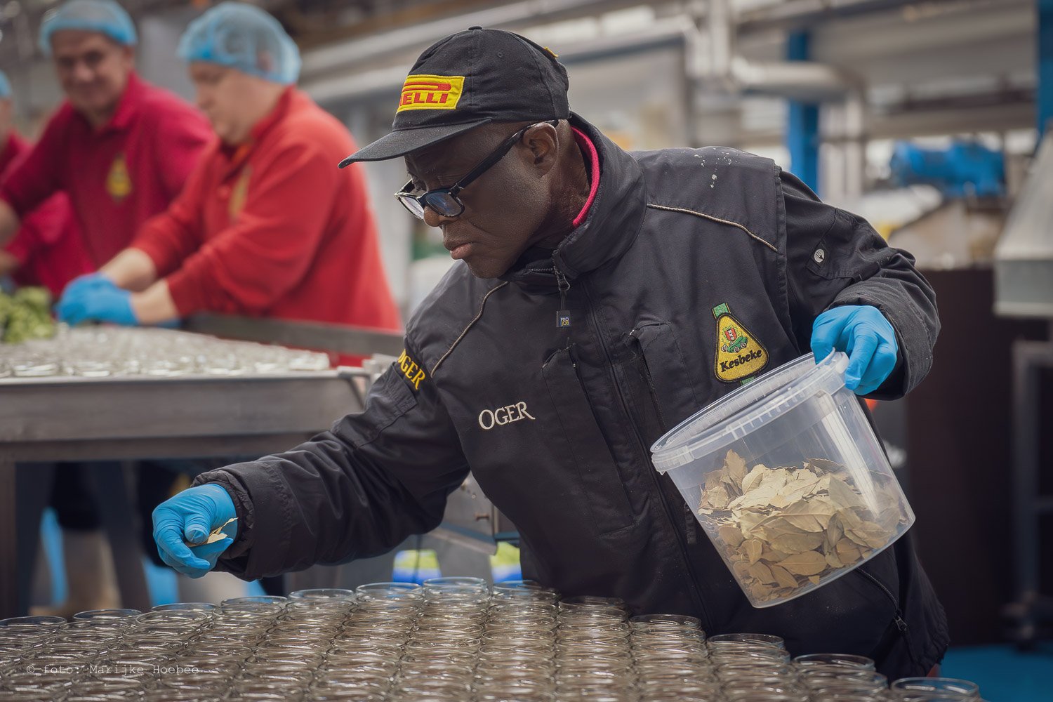 Man in een fabriek, met blauwe handschoenen, een zwarte jas en een pet met het Pirelli logo, vult kleine glazen potjes in met een witte stof.