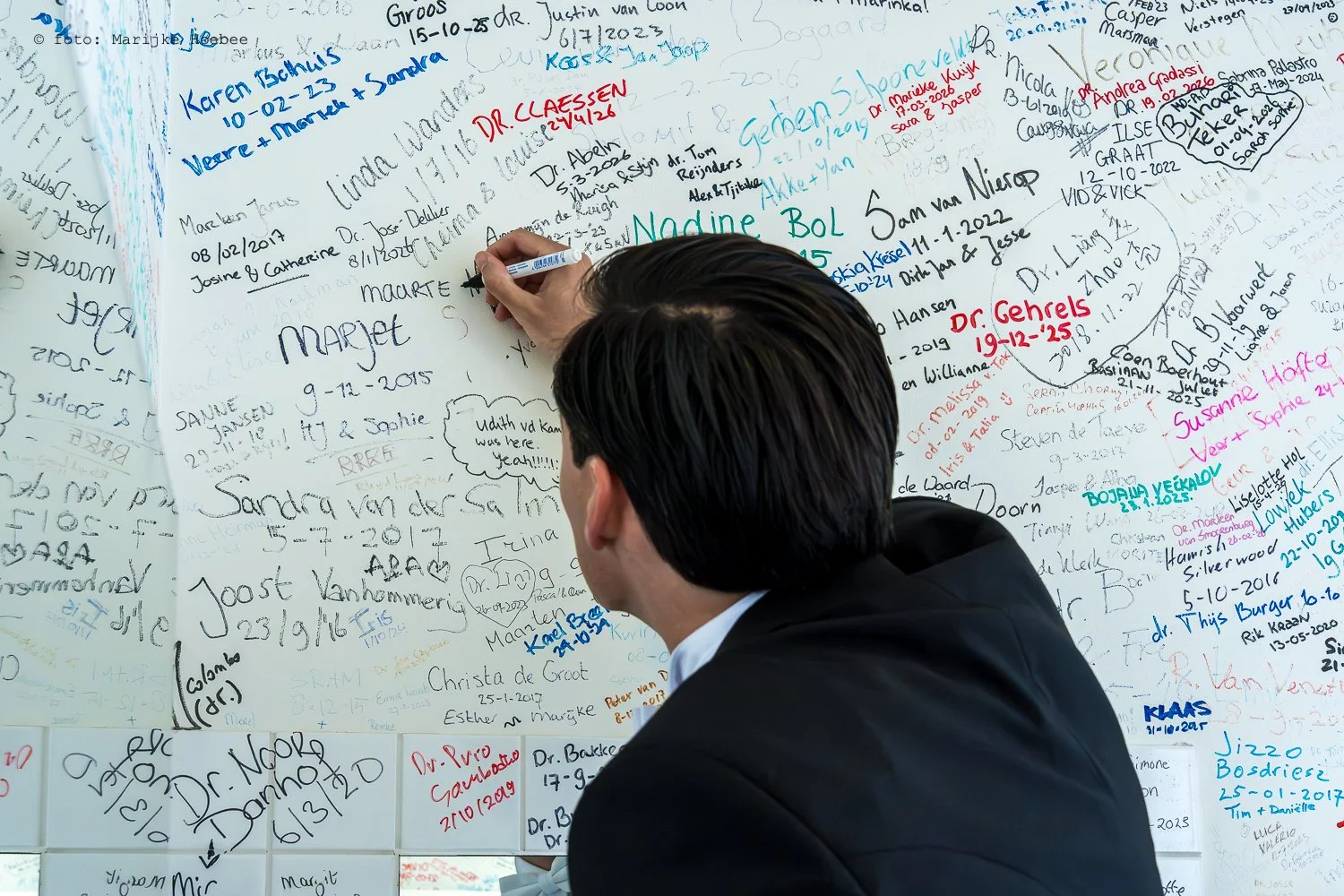 Een man in een zwart pak schrijft op een muur vol met handgeschreven namen en data in verschillende kleuren text.