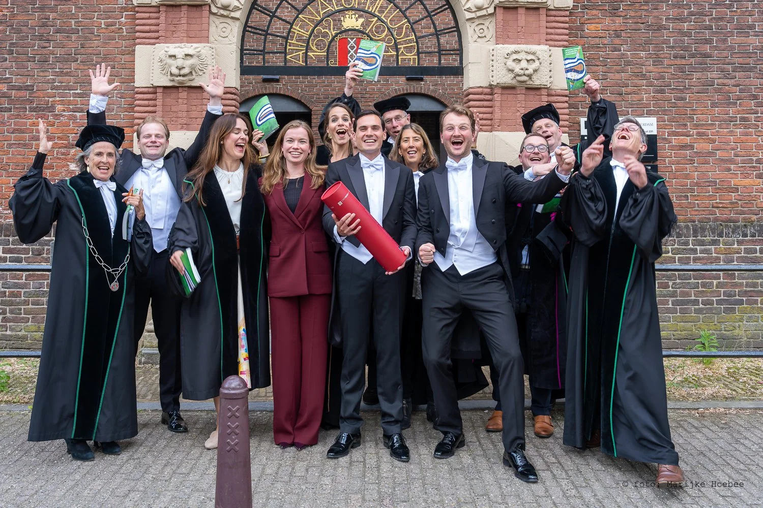 Groepsfoto van afgestudeerden in academische kleding voor een stenen gebouw met een ronde poort en lopende banden in de achtergrond, allemaal lachend en feestend.
