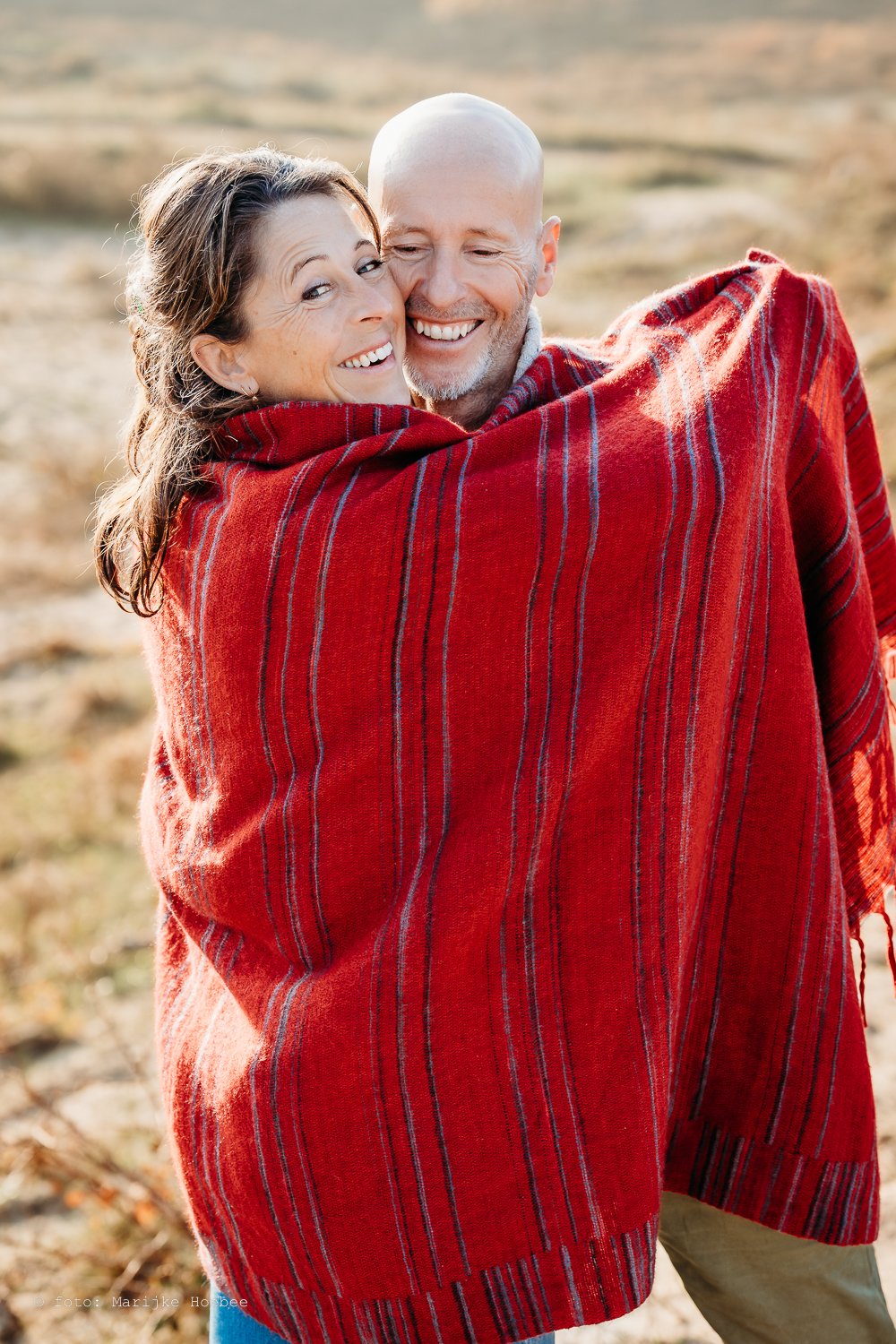 Twee mensen die samen lachen en omarmen, ingepakt in een rode shawl, buiten in een landelijke omgeving.
